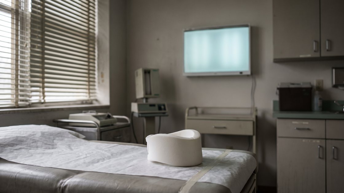 An empty hospital examination room with a cervical collar on the treatment table and an illuminated X-ray viewing panel