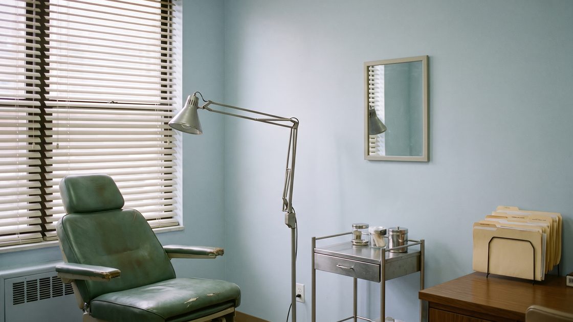 An empty medical consultation room with an examination chair, an articulated lamp and a rack of file folders on the desk