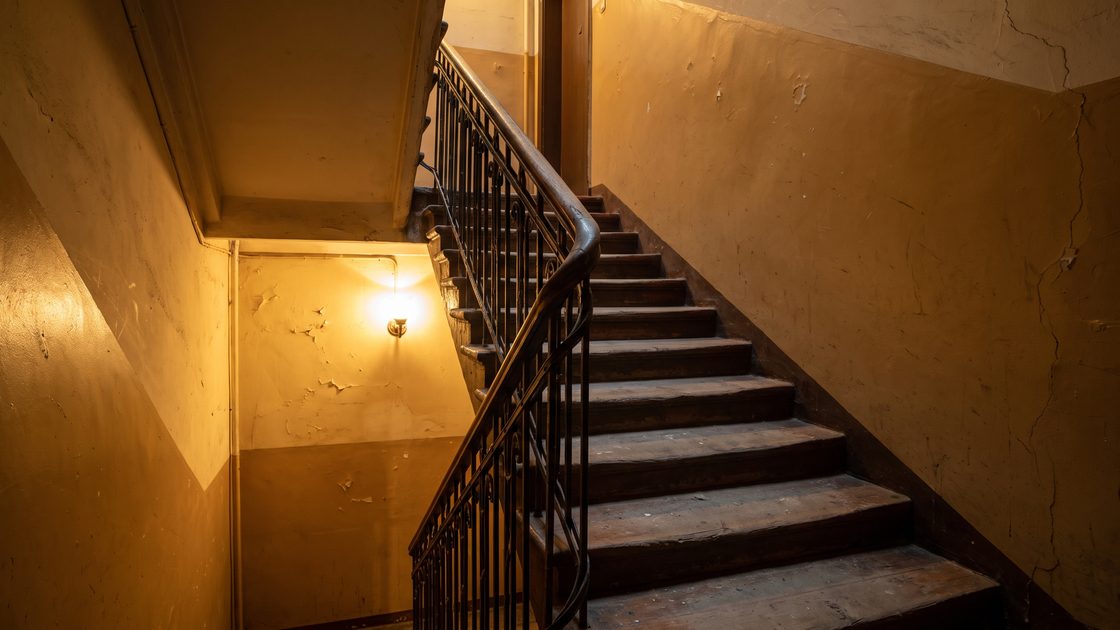 A dimly lit stairwell in an older apartment building with worn steps, a metal handrail and a single wall light