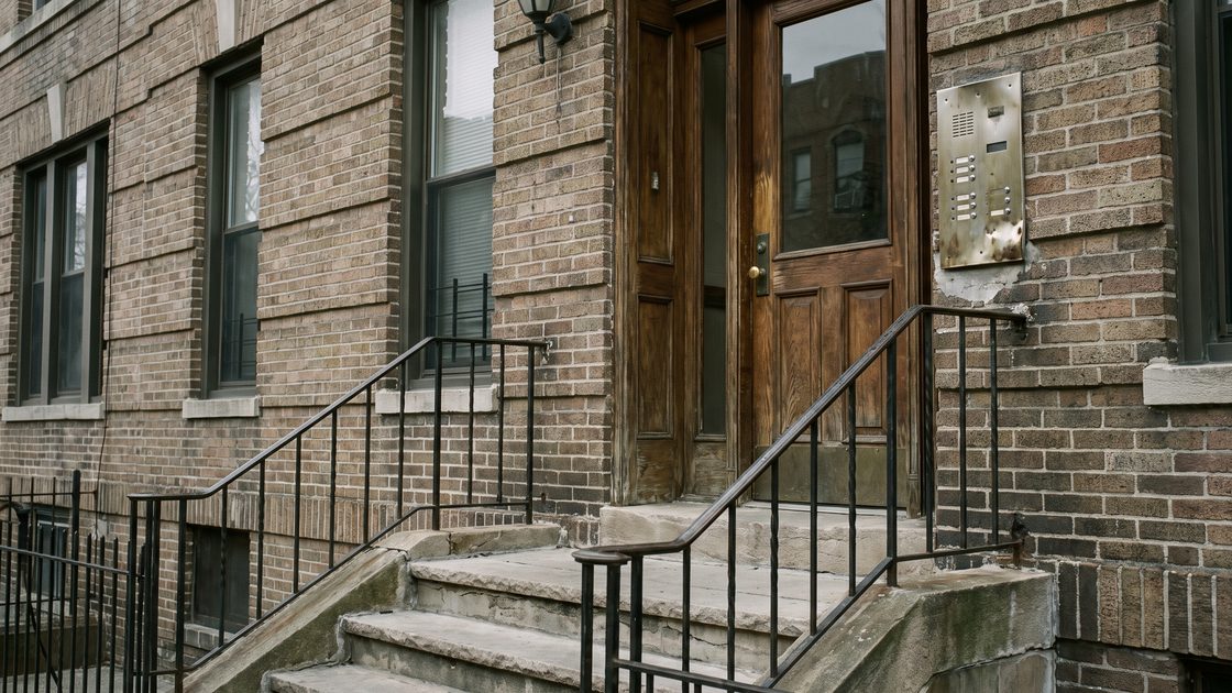 Stone steps and an iron railing at the entrance of a prewar brick apartment building, with an intercom panel beside the door