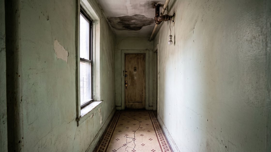 A hallway in an older apartment building with a water-stained ceiling and an exposed pipe