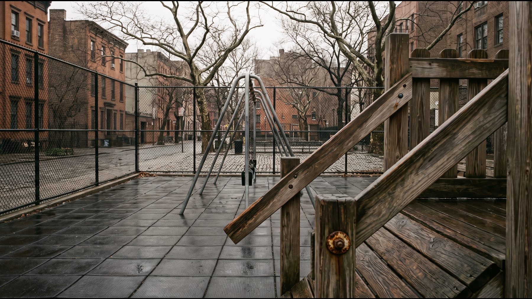 An empty New York City playground on an overcast morning with wet safety surfacing and a climbing frame
