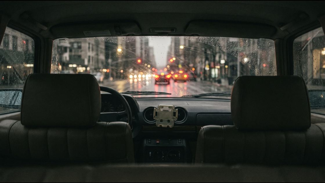 The view from the rear seat of a car at night looking through a rain-streaked windshield at blurred tail lights